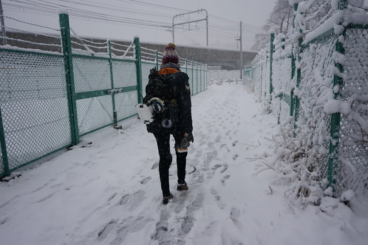 Je marche dans une rue enneigée d'une ville japonaise en costume d'agent de la SHD ; au loin on distingue un pont et des pilônes pour les trains de la ligne Chuo