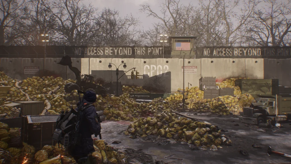 Une capture d'écran de la "Dark Zone", la zone PvP du jeu Tom Clancy's The Division ; des sacs de déchets contaminés jonchent le sol de cette rue de Manhattan barricadée par des blocs de ciment sur lesquels des panneaux proclament "no access beyond this point", bloquant l'accès à Central Park